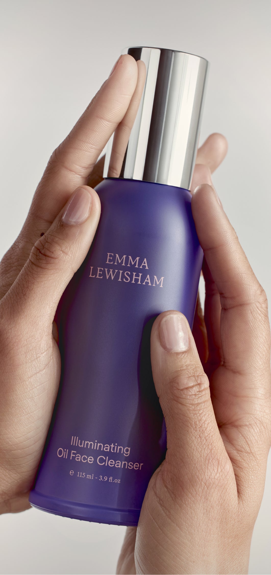 Hand holding a purple bottle of Emma Lewisham illuminating oil face cleanser against a neutral background